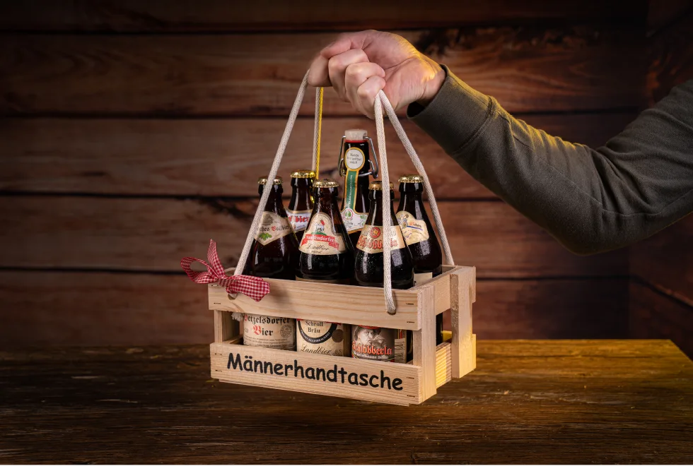 Eine Hand hält ein Holztragerl mit der Aufschrift "Männerhandtasche" in dem 6 Flaschen Bier sind